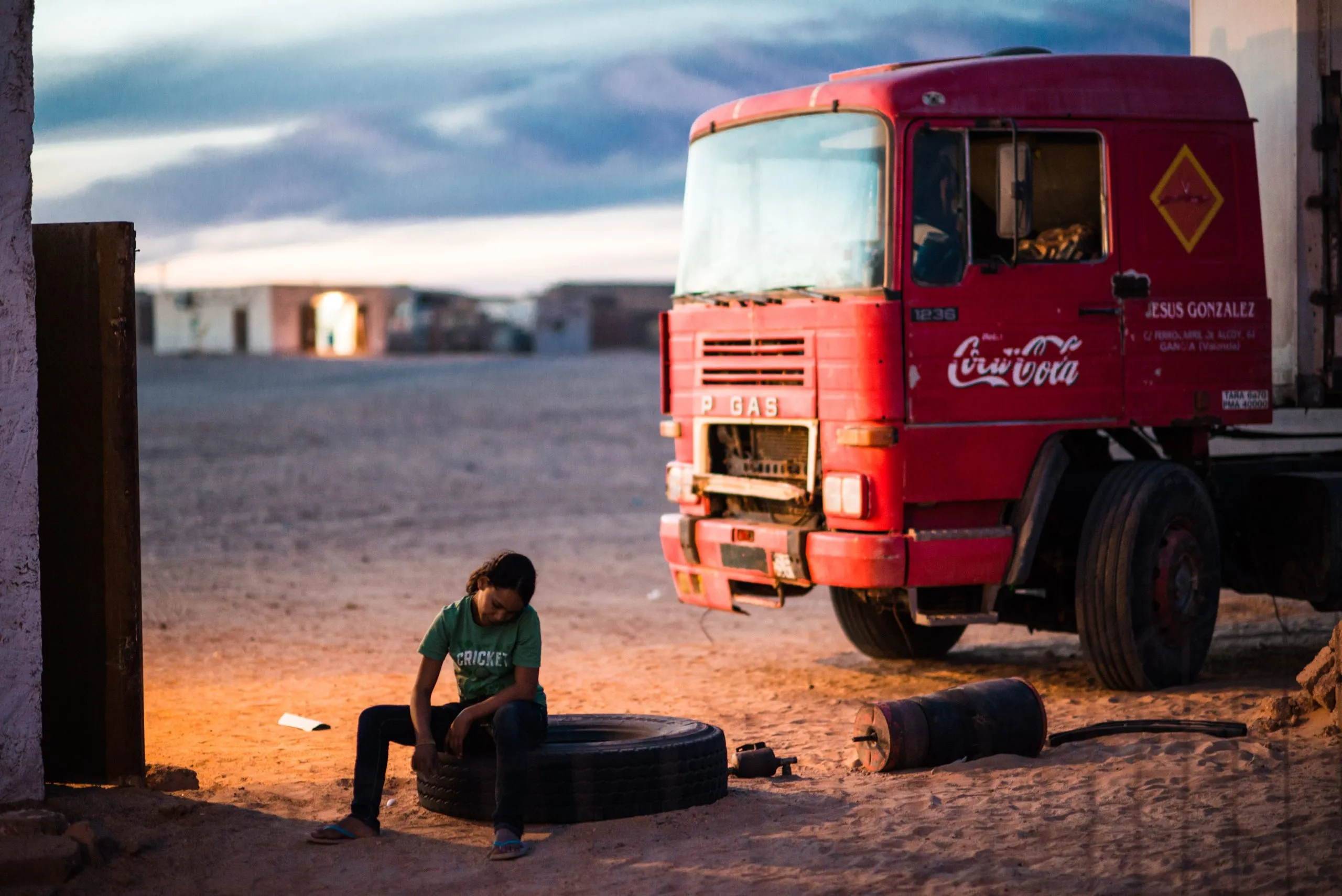A girl reads a book seated on a stack of tyres beside an old red truck at dusk in the Tindouf camp.