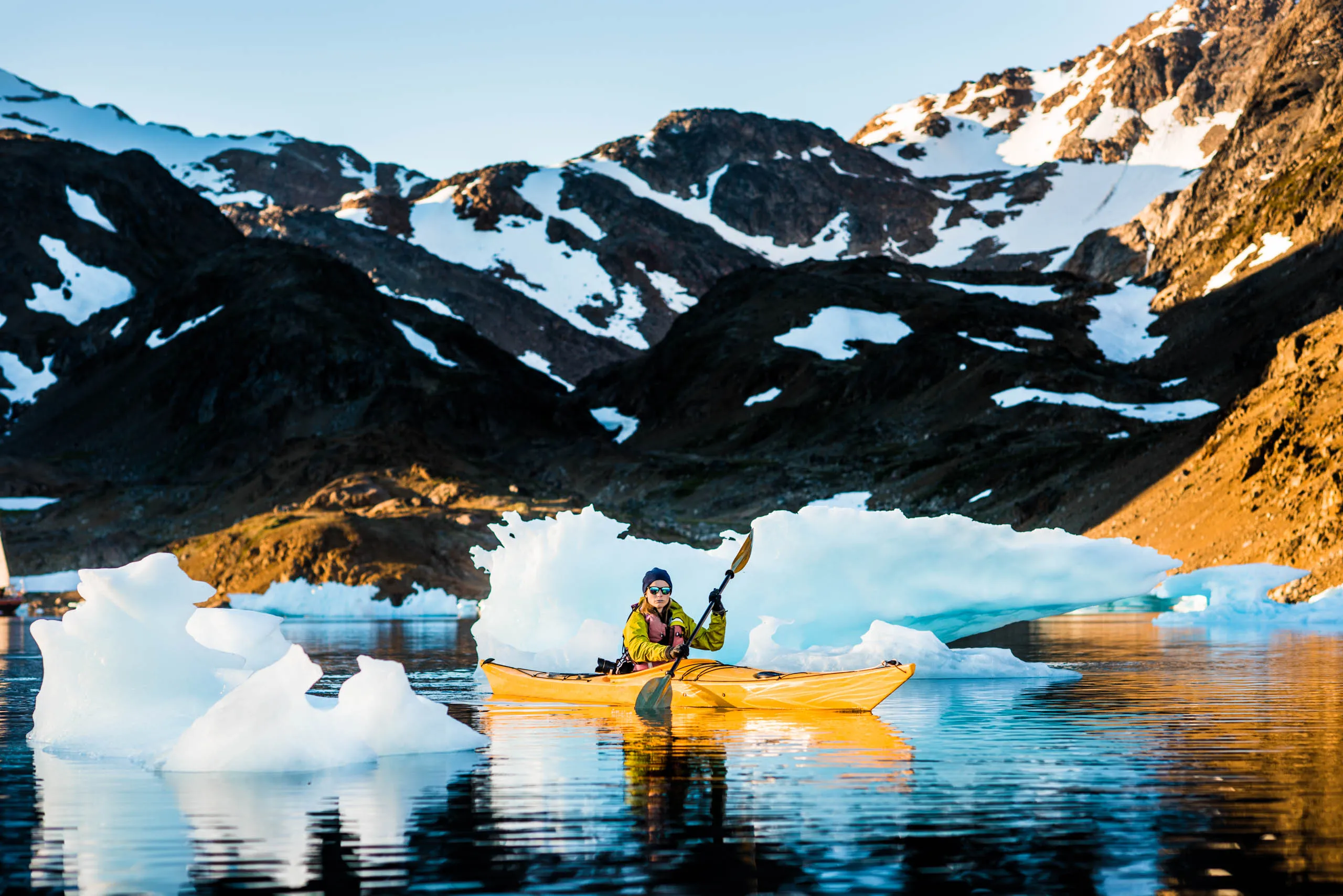 Kayaker in yellow boat paddling among small icebergs below snow-streaked Greenland mountains