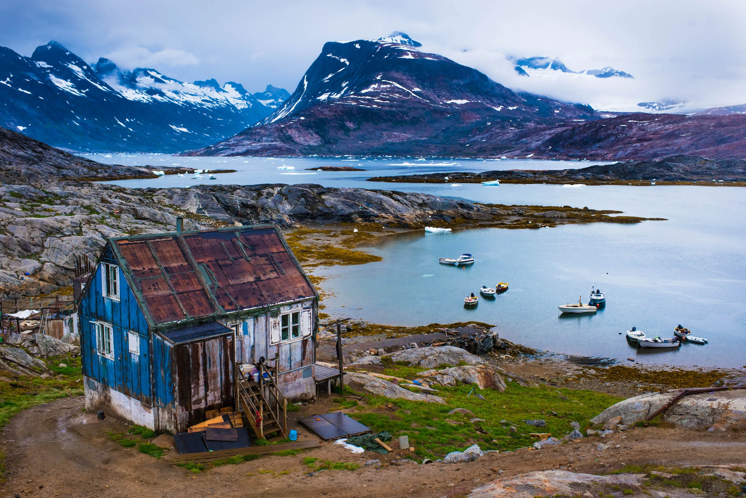 Weathered blue cabin beside a calm fjord with snow-capped peaks and anchored boats