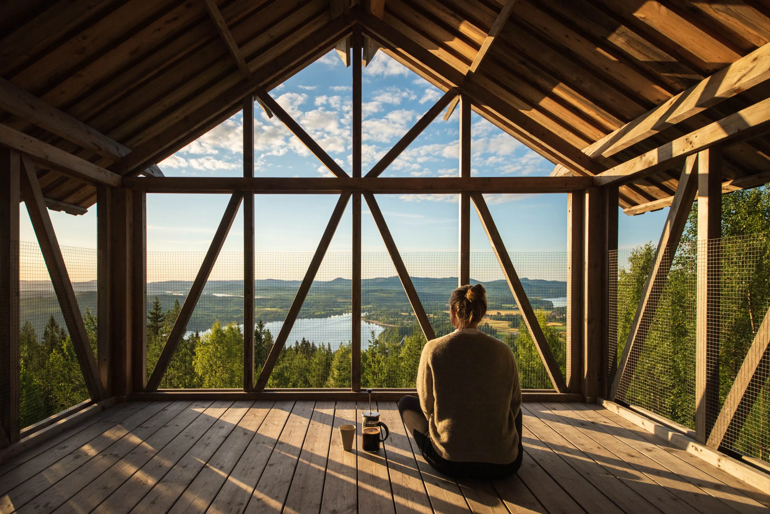 A woman sits on the floor of an open loft in the morning light, overlooking the mountains and forest of Orbaden in Hälsingland.