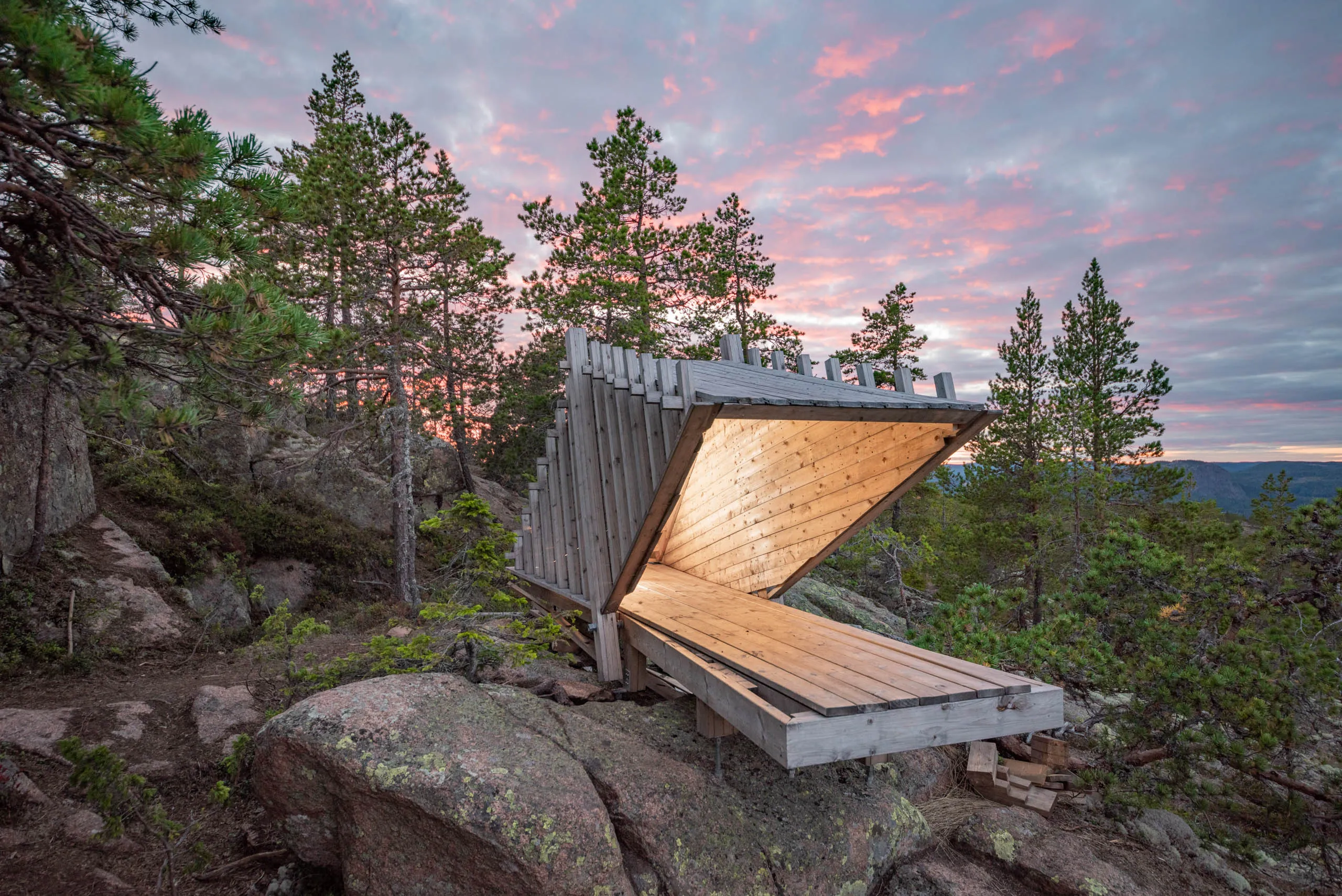 A hut built with minimalistic architecture stands on top of a forested hill of the High Coast of Sweden, with a colorful twilight sky behind it.